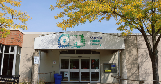 Image of an Oakville Public Library Entrance - a white arhc in the Centre shows the library's logo, with sliding doors seen below the arch. At the top of the image is a blue sky and trees are on either side.