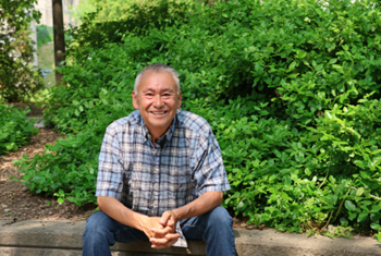 Stephen-Webpage-Banner.png Image of a man smiling and sitting down. Behind him are green bushes.