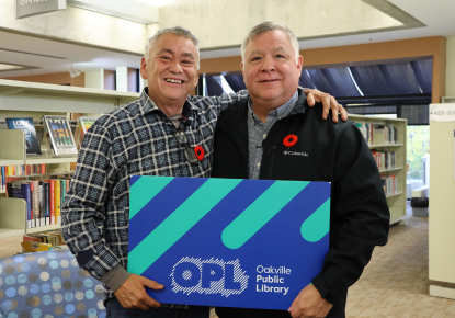 Image of two men smiling and holding a large Oakville Public Library card that is blue and teal with a white logo. In the background are bookshelves.