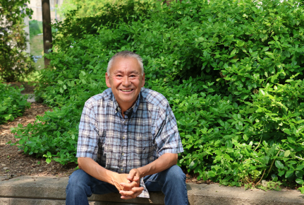 Image of a man smiling and sitting down. Behind him are green bushes.